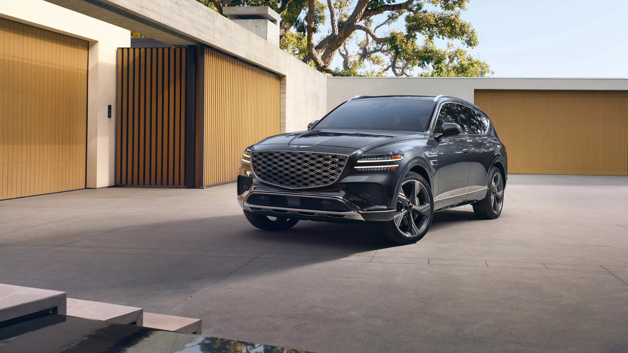 3/4 front driver side view of Genesis GV80 in Makalu Gray parked in driveway of modern house