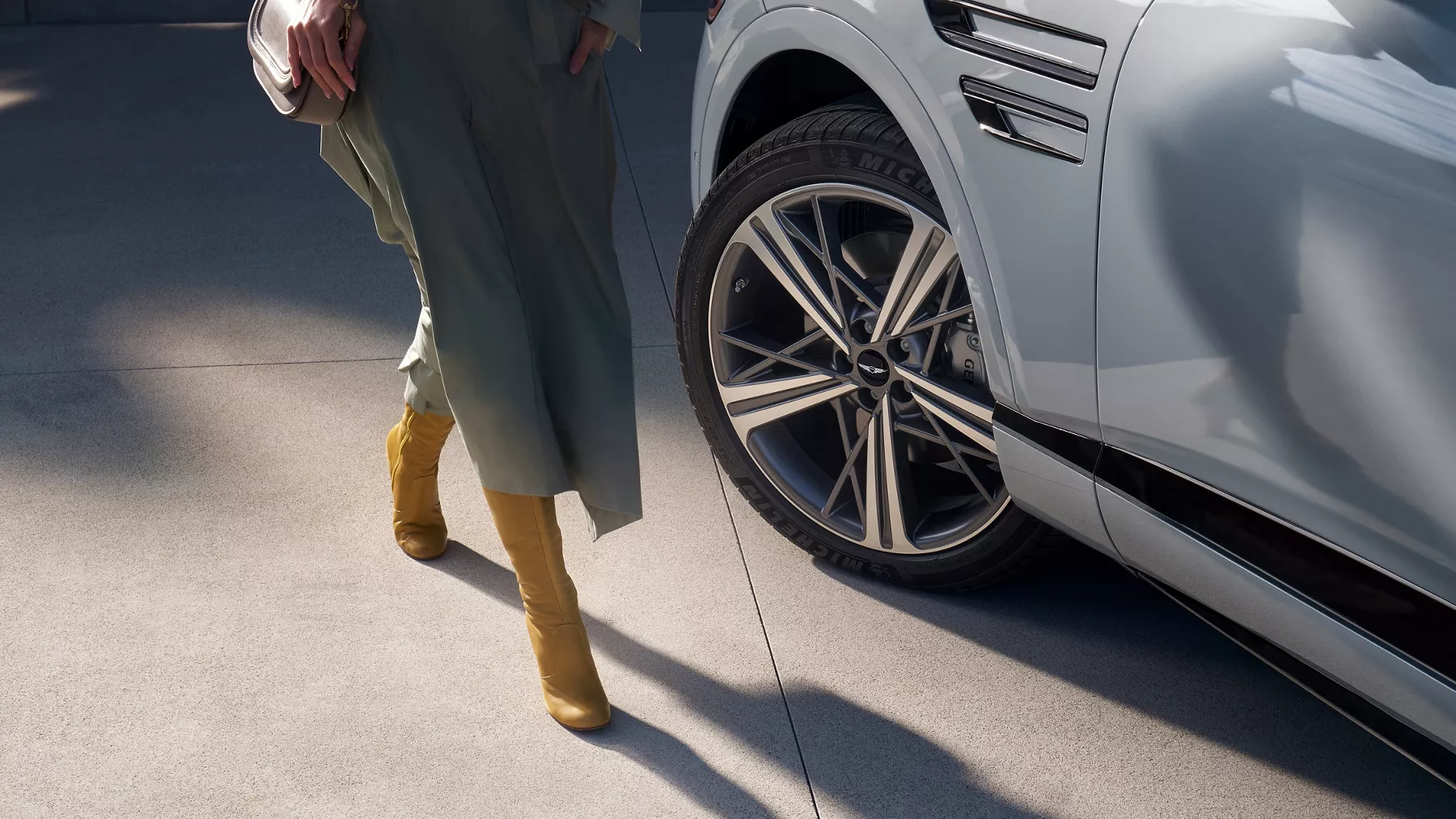 Close up of 20-inch dark gray matte rims on Genesis GV80 Coupe in Bering Blue, woman in dress nearby