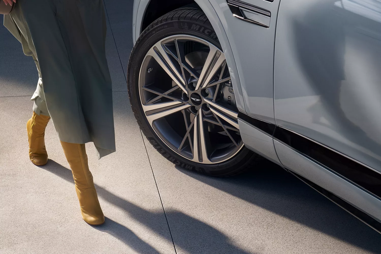 Close up of 20-inch dark gray matte rims on Genesis GV80 Coupe in Bering Blue, woman in dress nearby