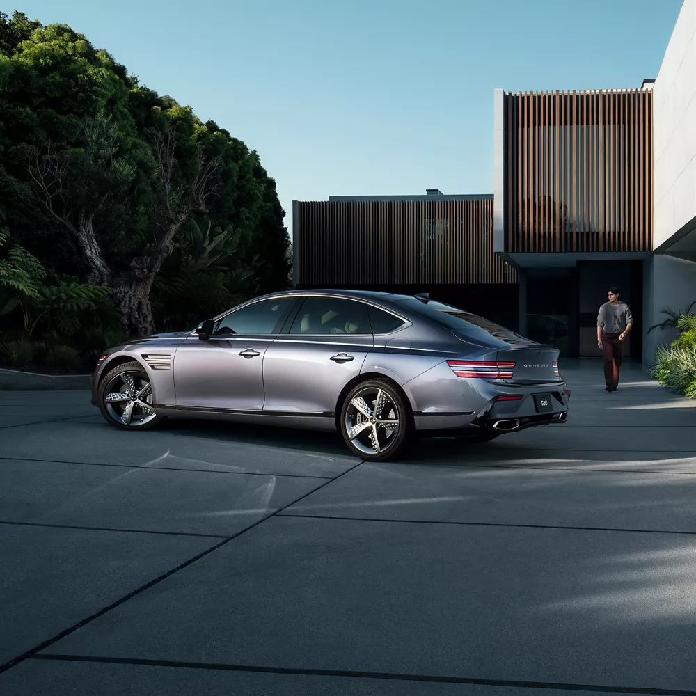 Driver side profile view of Genesis G80 in Ceres Blue parked next to modern house with man nearby