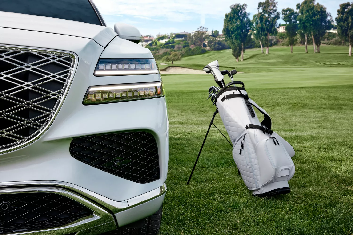 Close up of grill and headlight on white Genesis GV80 next to a white golf bag on a golf course