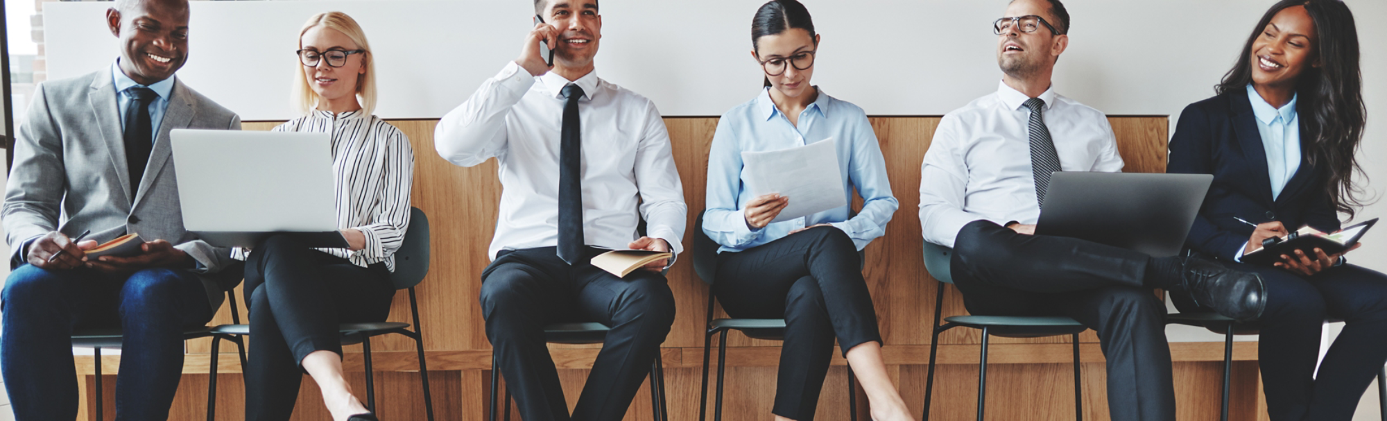 Group of diverse businesspeople smiling while sitting in a row in an office reception waiting for a meeting