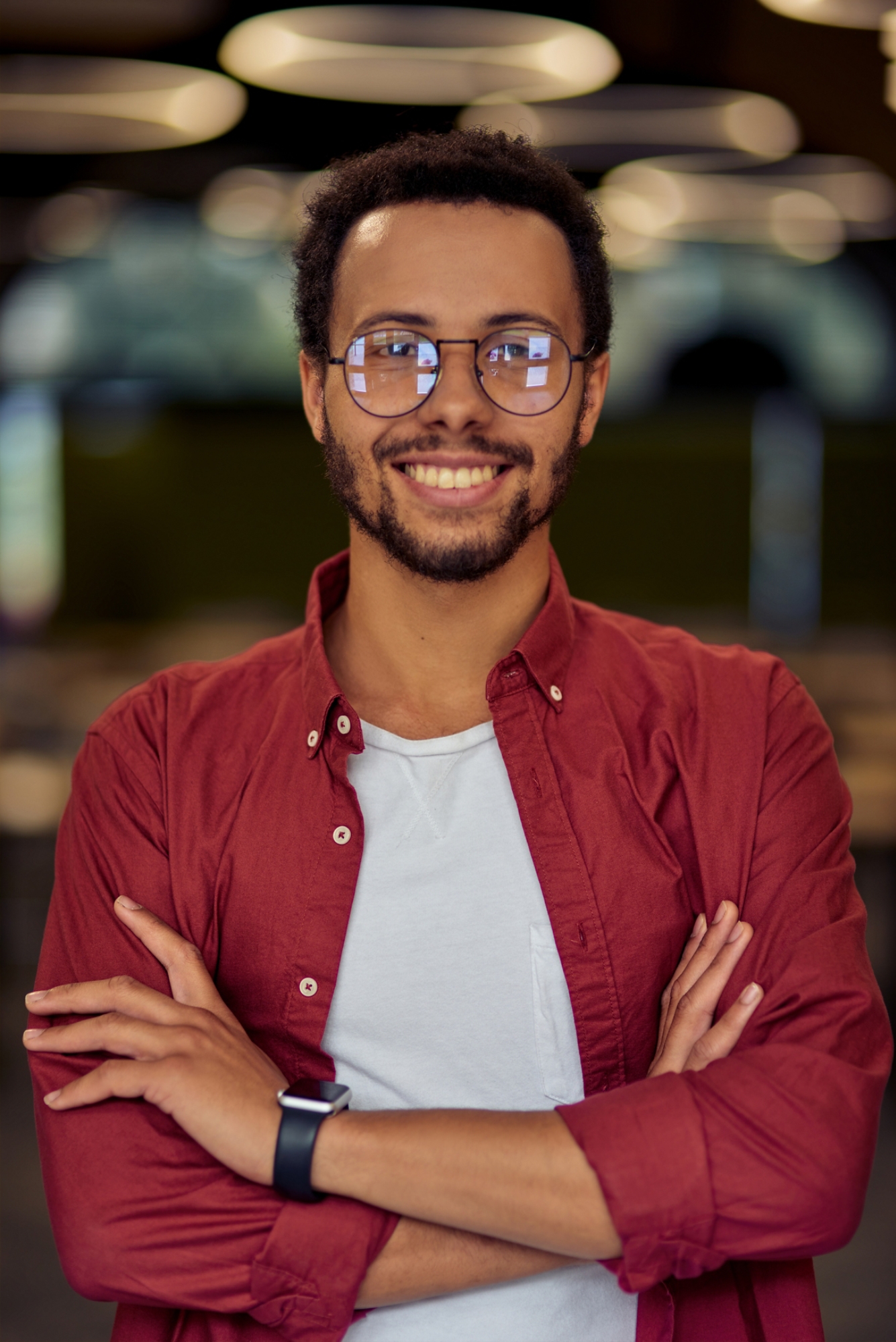 Vertical shot of a young happy mixed race businessman or office worker in casual wear keeping arms crossed, looking at camera and smiling while posing in the office. Business people concept