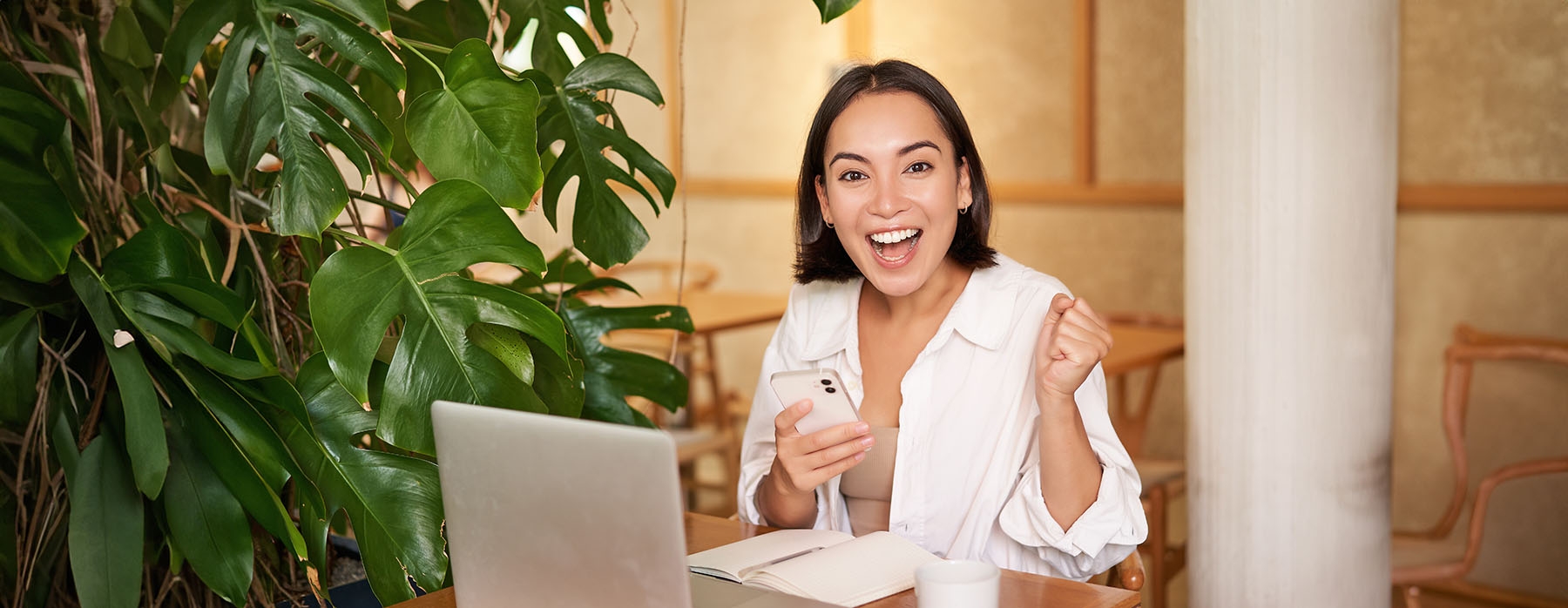 Joyful young asian woman winning, celebrating victory or achievement, receive good news on mobile phone, sitting with laptop and working.