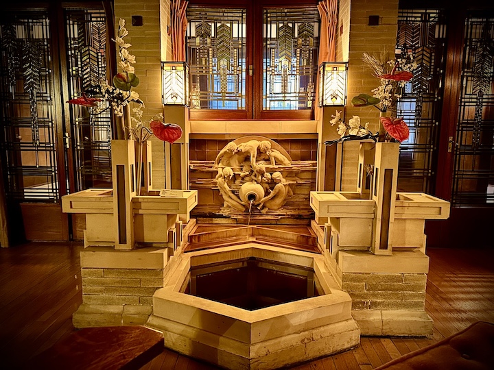 Moonchildren fountain at Dana-Thomas House. Concrete water fountain inside a building featuring a sculpture of children gathered around an urn, pouring water into a hexagonal pool. Concrete flower boxes on each side of fountain; geometric stained glass windows and doors behind fountain..