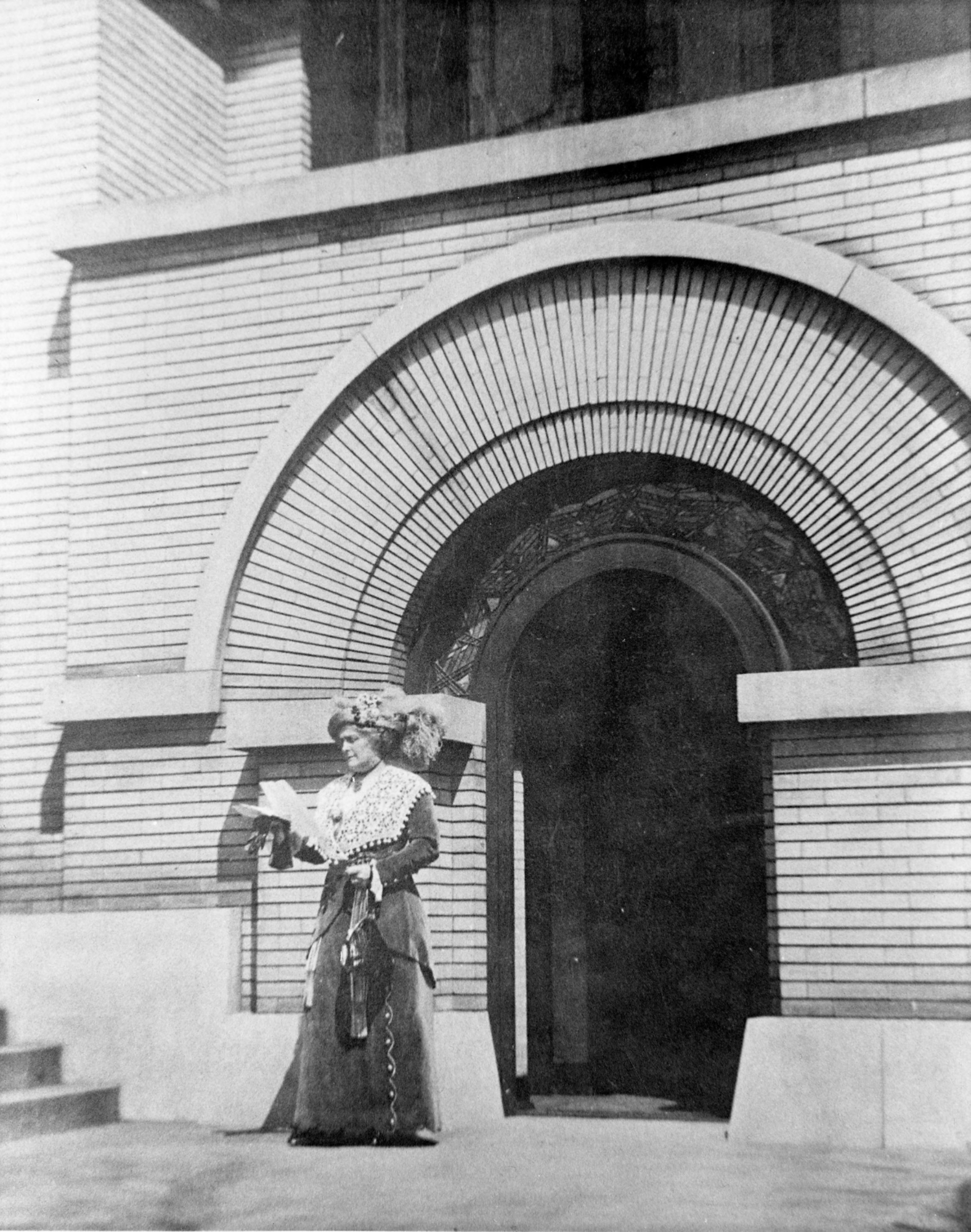 An early 20th century woman (Susan Lawrence Dana)  stands outside the door of a building reading a document.