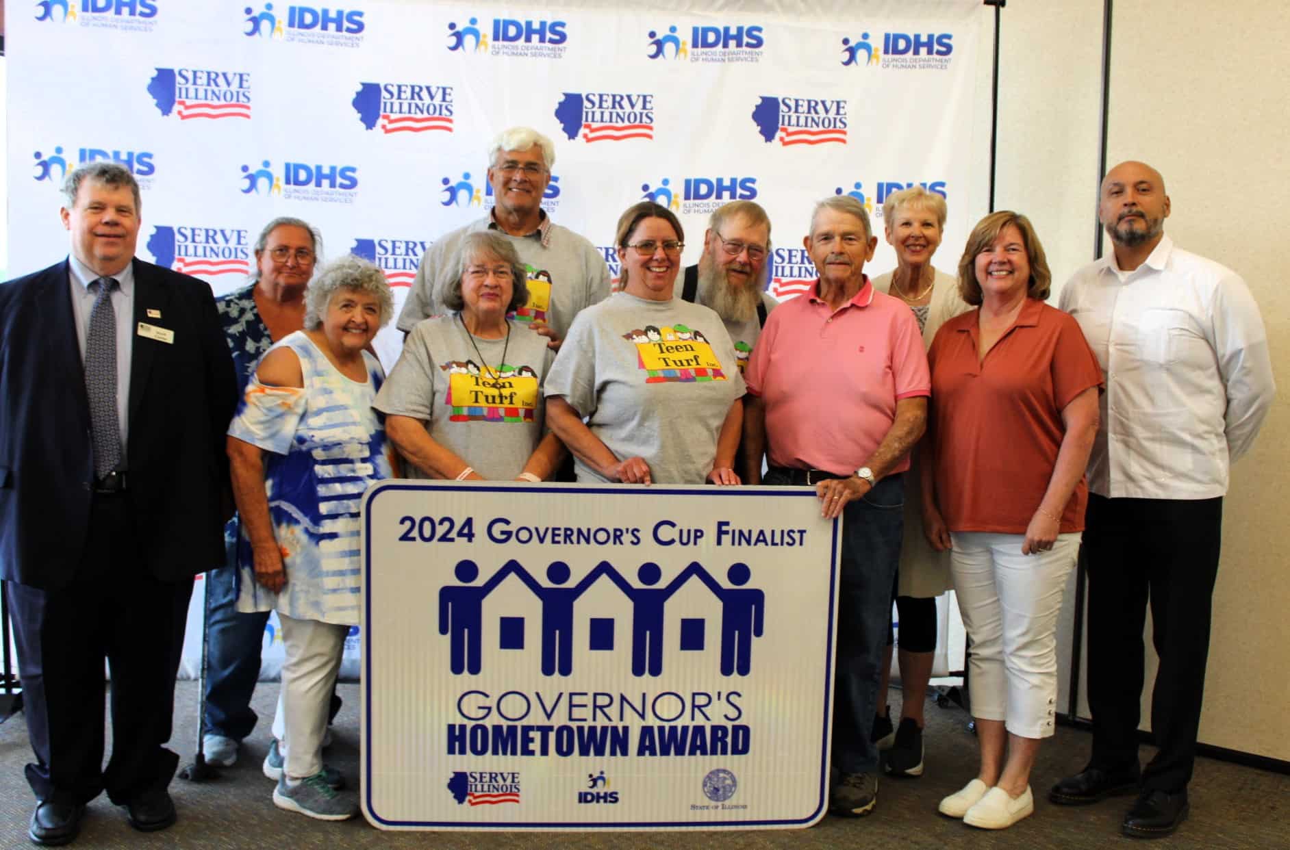 Group photo of Teen Turf representatives, Commissioners, and Serve Illinois Executive Director Andres Fernandez with Governor's Cup Finalists highway sign