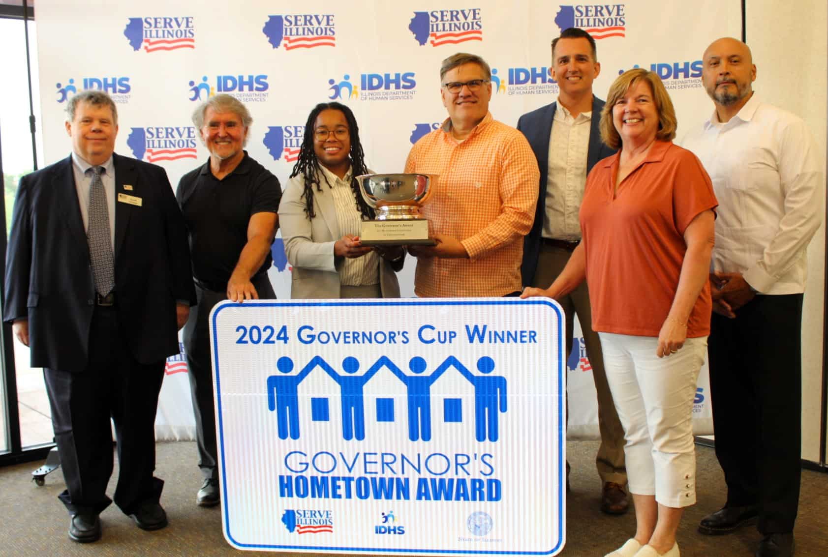 Group photo of Challenger Baseball representatives, Commissioners, and Serve Illinois Executive Director Andres Fernandez with Governor's Cup winner highway sign