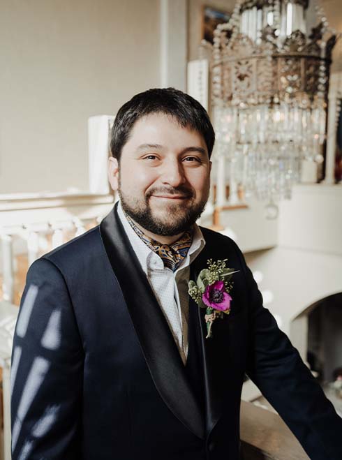 D. Javier Thompson, a Latine nonbinary adult with short dark brown hair and brown eyes, smiles at the camera. Javier is wearing a dark blue tuxedo with black collared jacket, white collared shirt, blue plus gold paisley ascot, and green and pink boutonniere pinned to their right tuxedo jacket pocket. Behind them is a staircase and crystal chandelier. 