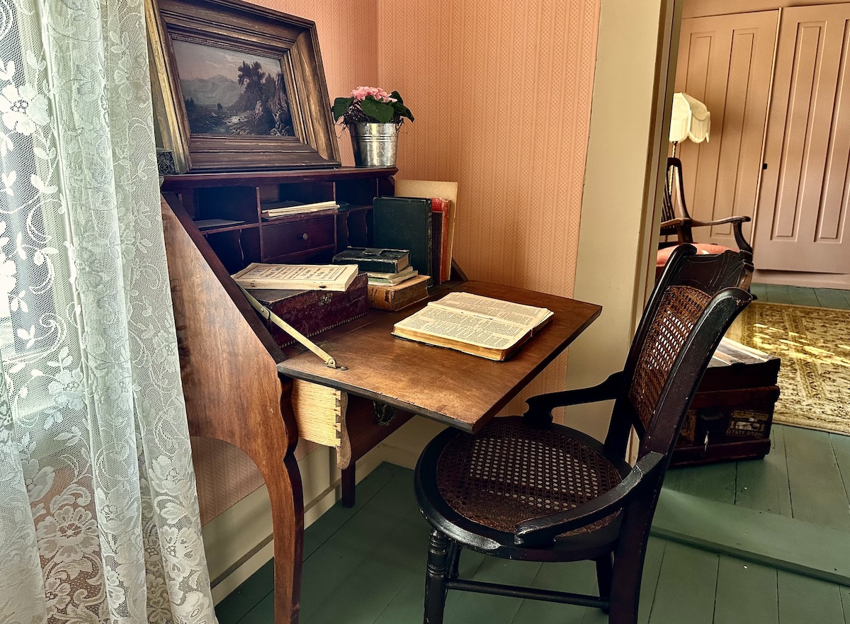 A nineteenth century desk with books, papers and personal items scattered on it. A nineteenth century wooden chair is pushed in under the desk