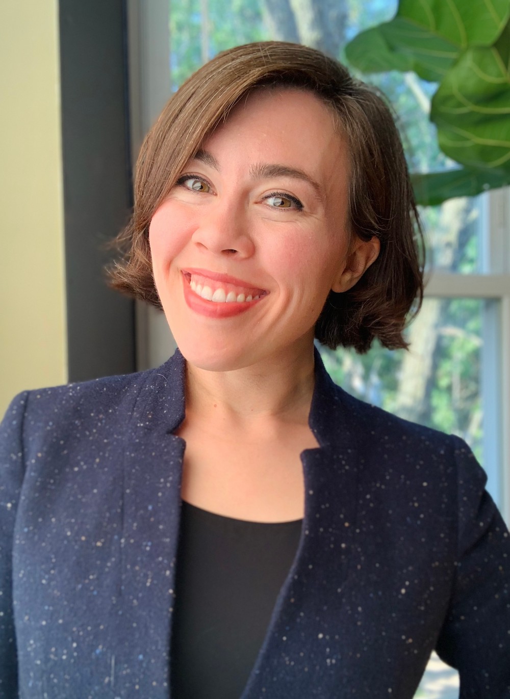 Dr. Addie Shrodes, a white femme with chin-length brown hair and brown eyes, smiles as they look at the camera. They are wearing a navy speckled blazer and a black shirt. Behind her is a fiddle leaf fig tree leaf and a window overlooking trees.