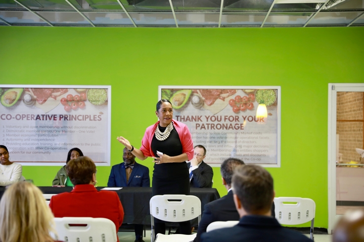 Candid photo of Lt. Governor Stratton speaking to a small group of people, we see the back of their heads, and three people seated at a table with a black table cloth behind her. She is wearing a black dress, pink shawl, and pearl necklace. Her hair is pulled back and she appears to be mid-sentence, with her left hand raised at her waist. There is a bright green wall in the background with framed signs. One of the signs reads “Thank you for your patronage.” There are vegetables on the signs. 