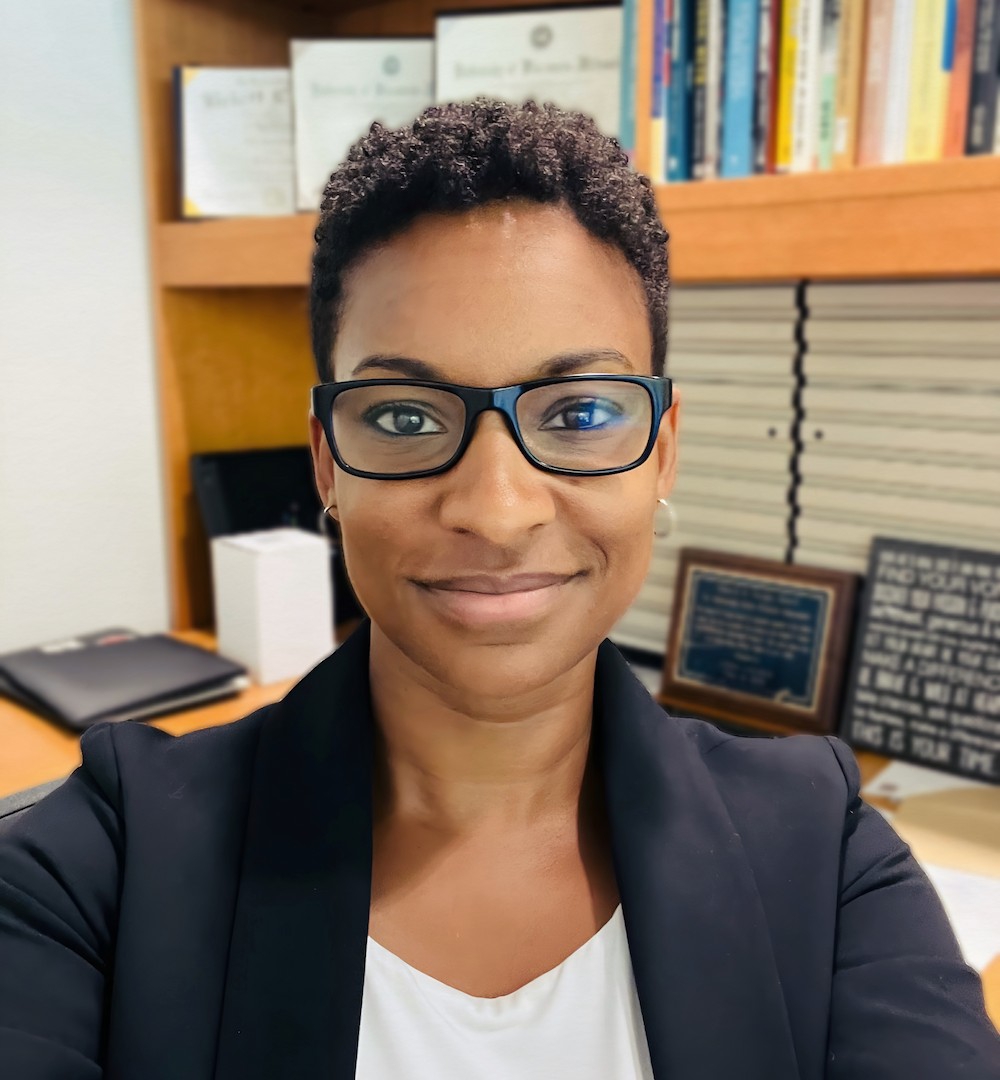 Dr. Atiera Coleman, a Black woman with short curly black hair, brown eyes, and black glasses, smiles at the camera. Dr. Coleman is wearing a white blouse and a black blazer. Behind her is a desk and a bookshelf filled with books and diplomas.