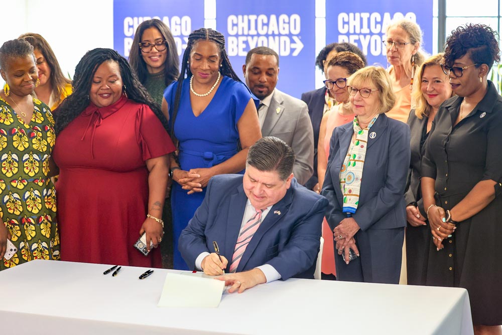 Gov. Pritzker, seated at a table, signs the Birth Equity Initiative bill with Lt. Governor Stratton and others smiling, standing behind him: Chicago South Side Birth Center founder Jeanine Valrie Logan, Sen. Lakeisha Collins, Personal PAC President & CEO Sarah Garza Resnick, Chicago Beyond CEO Liz Dozier, Rep. Anna Moeller, Rep. Mary Beth Canty, Rep. Diane Blair-Sherlock, Sen. Willie Preston, Sen. Adriane Johnson, Leader Robyn Gabel, and Rep. Yolonda Morris.