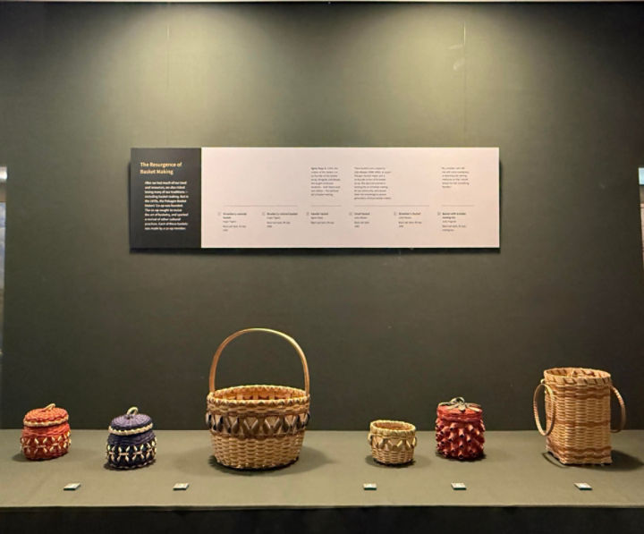 Six black ash baskets sitting horizontally on a green display shelf. An exhibit label is on the back wall.