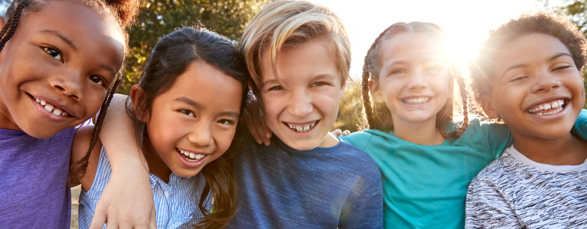 Five children, a mix of boys and girls, smiling with their arms draped over each other’s shoulders outside on a sunny day.