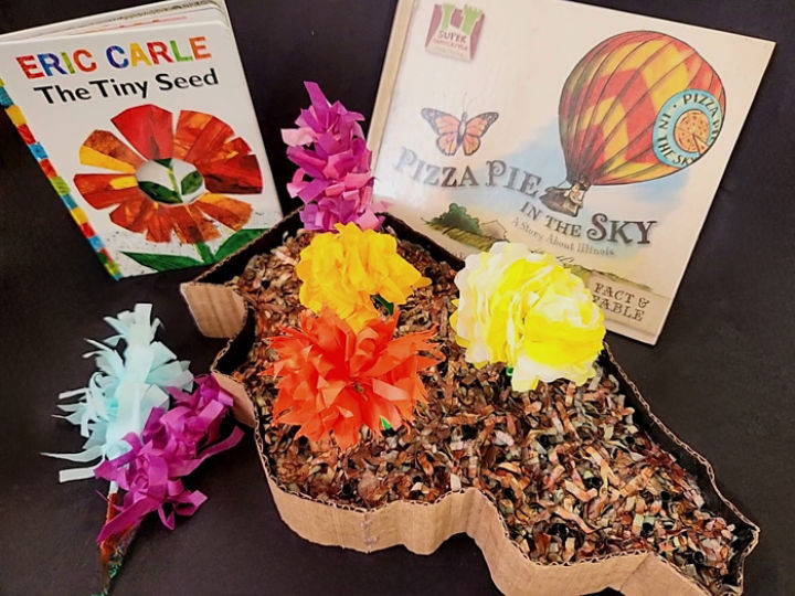 Two books stand up next to each other in front of a black backdrop. Sitting in front of the books, an Illinois shaped flower bed craft made from cardboard and shredded paper holds purple, yellow, and orange tissue paper flowers.