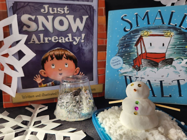 Two books picturing a child and a snow play stand up behind paper snowflakes. In front of the books a small snowman sits atop a pile of fake snow next to a snow globe with a miniature mammoth inside. 