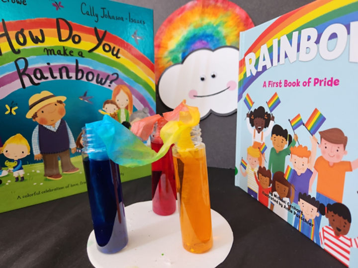 Two children’s books about rainbows surround a rainbow coffee filter and smiling cloud craft. In front of the books, test tubes containing yellow, blue, and red water are linked to make a rainbow.