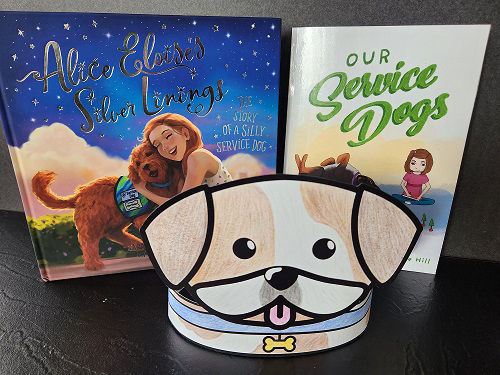  A paper cut out of a colored brown and white dog with a blue collar sits in front of two books about service dogs. The cover of the left book, titled Alice Eloise’s Silver Linings, features a woman hugging her double doodle puppy who wears a green service dog vest. The cover of the other book, Our Service Dogs, shows a girl sitting cross legged on the floor with her dark brown dog laying on its back next to her and a purple service dog vest placed in front of them.