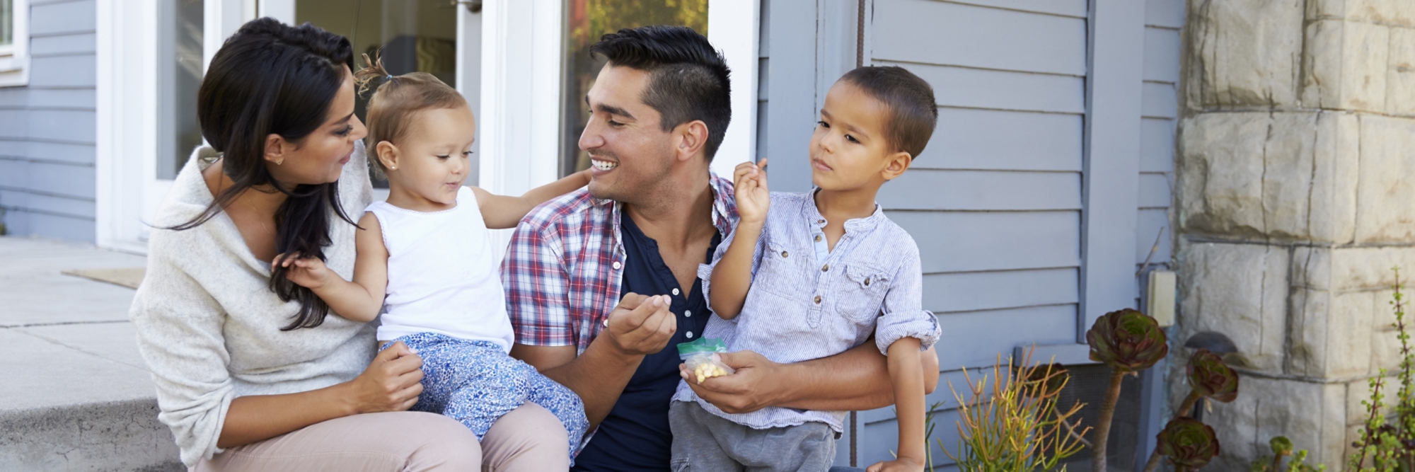 Family sitting together on the steps outside their house, with the young daughter on the mother’s lap and the young son on the father’s lap.
