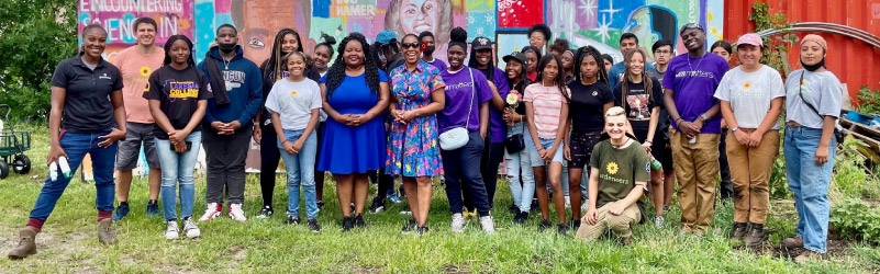 Lt. Governor Stratton poses for a group photo with a group of Black and Brown youth wearing jeans and t-shirts. They are outside, standing in a grassy area. There is a colorful mural slightly blurred in the background. She is wearing a short sleeve floral dress and sunglasses. Everyone is smiling. 