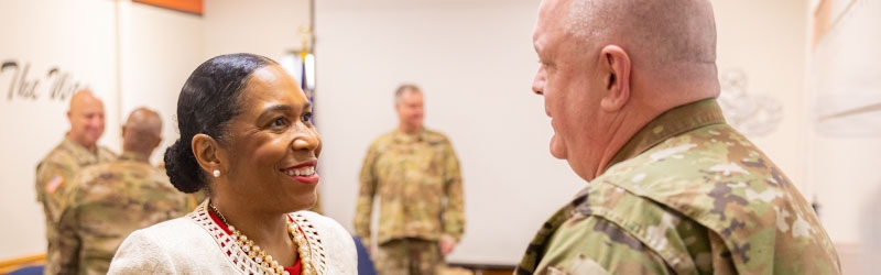 Candid photo of Lt. Governor Stratton, left, with a man in a camouflage uniform, right. The photo is cropped from the neck up. She is wearing a white blazer and a pearl necklace and earrings, smiling. We see the back and side of the military member’s head. In the background are more men in uniform. The background is blurred. 