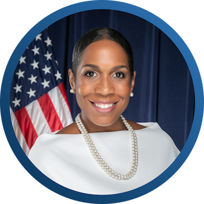 Headshot of Lt. Governor Stratton. The photo is a small circle crop thumbnail with a blue border. She is facing the camera wearing a white shirt, a pearl necklace and earrings. Her hair is pulled back with an American flag over her right shoulder. 