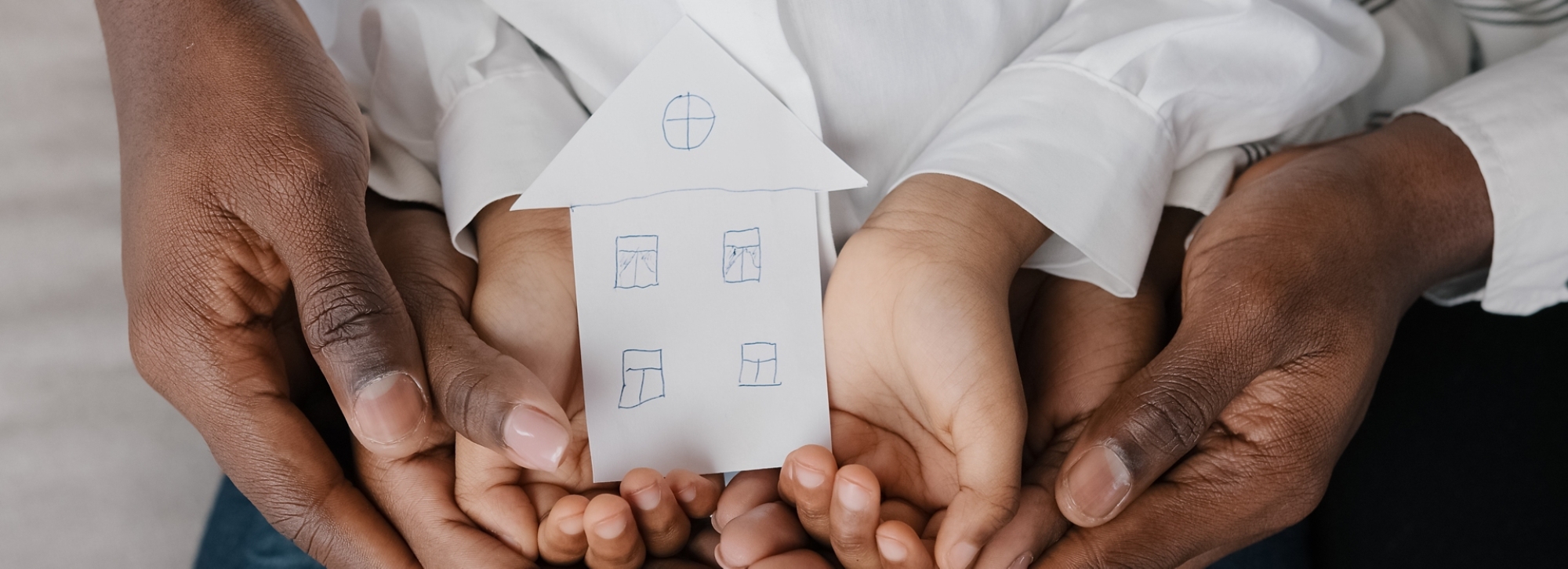 Close-up of an adult man’s hands cupping an adult woman’s hands, which are in turn cupping a child’s hands which are holding a cardboard cutout of a house.