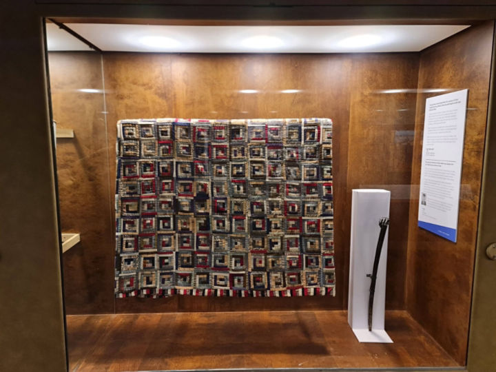 Image of exhibit case with a multicolor quilt and a dark wooden walking cane.