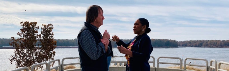 Candid photo of Lt. Governor Stratton, right, with a man wearing a long sleeved shirt and vest. The photo is cropped, and we are looking at their profiles, as they have a conversation. In the background is a body of water with some guardrails and a small tree in the left side of the frame.  