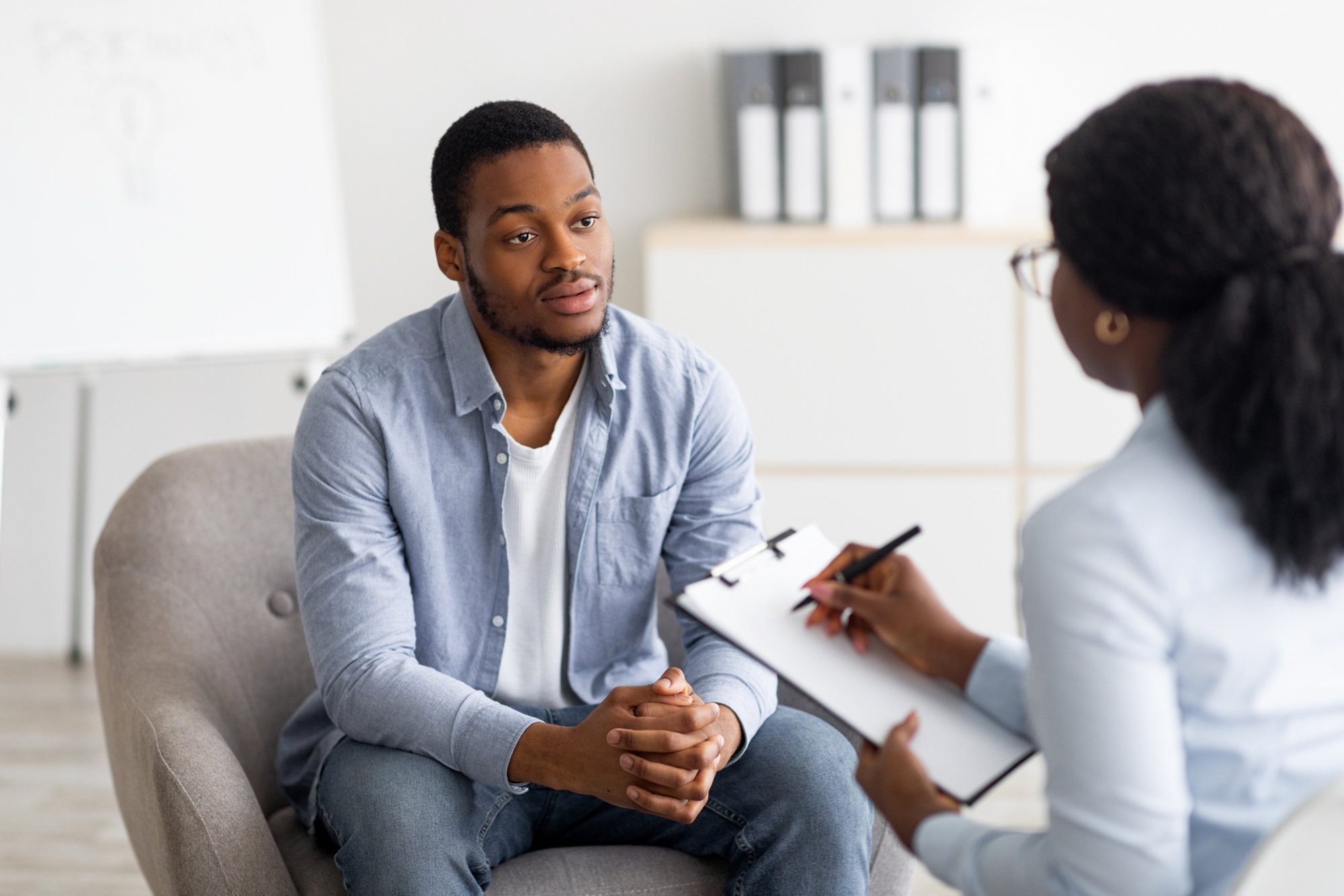 Unhappy young black man having session with professional psychologist at mental health clinic. Psychotherapist taking notes during conversation with depressed male patient; Shutterstock ID 1996252295; purchase_order: 19265; job: MHP_Landing_Page_Photos; client: Illinois Department of Insurance; other: 