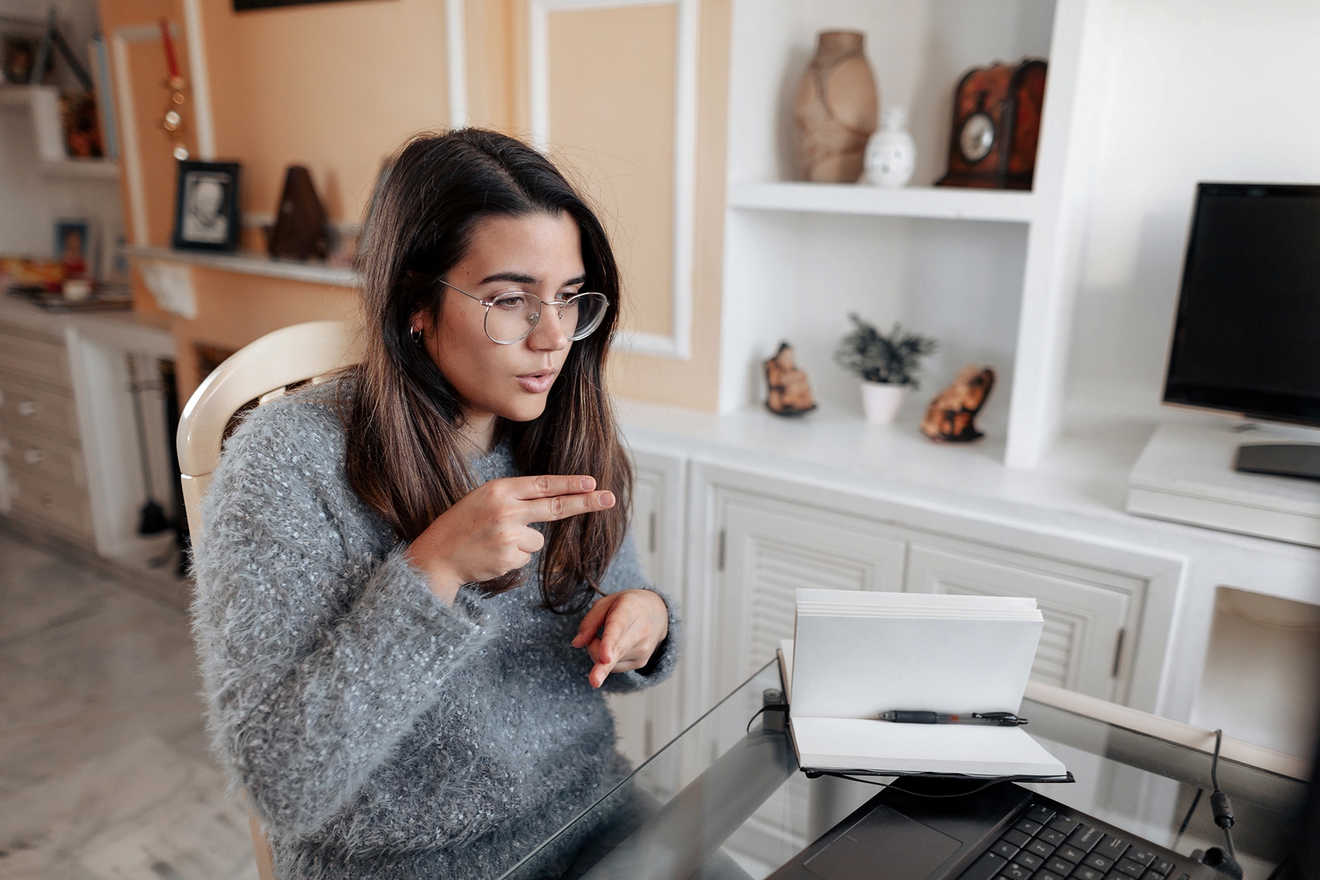 Medium shot young deaf woman wearing glasses and a sweater is using sign language to spell name while in an online video call in her living room.