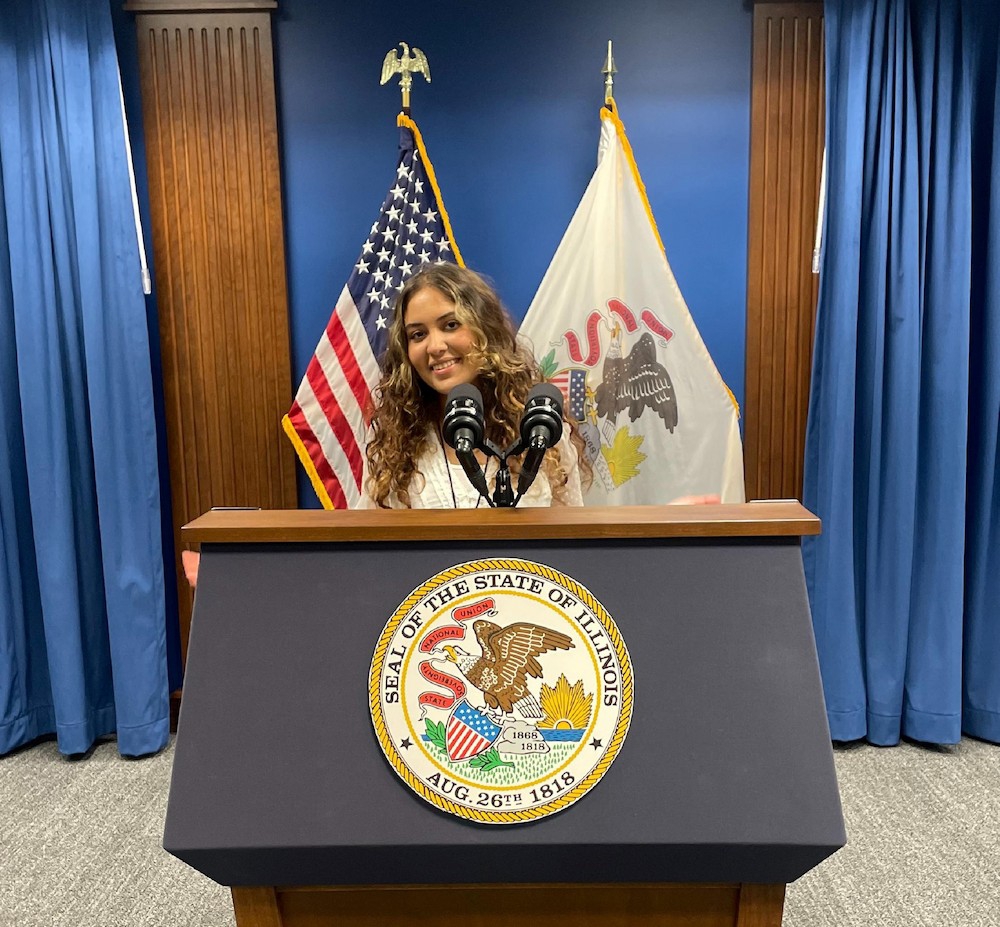 Headshot of Yosor Alwan. She stands behind a podium that bears the seal of the State of Illinois. She has long curly hair and wears a white blouse. Behind her are the United States and State of Illinois flags.