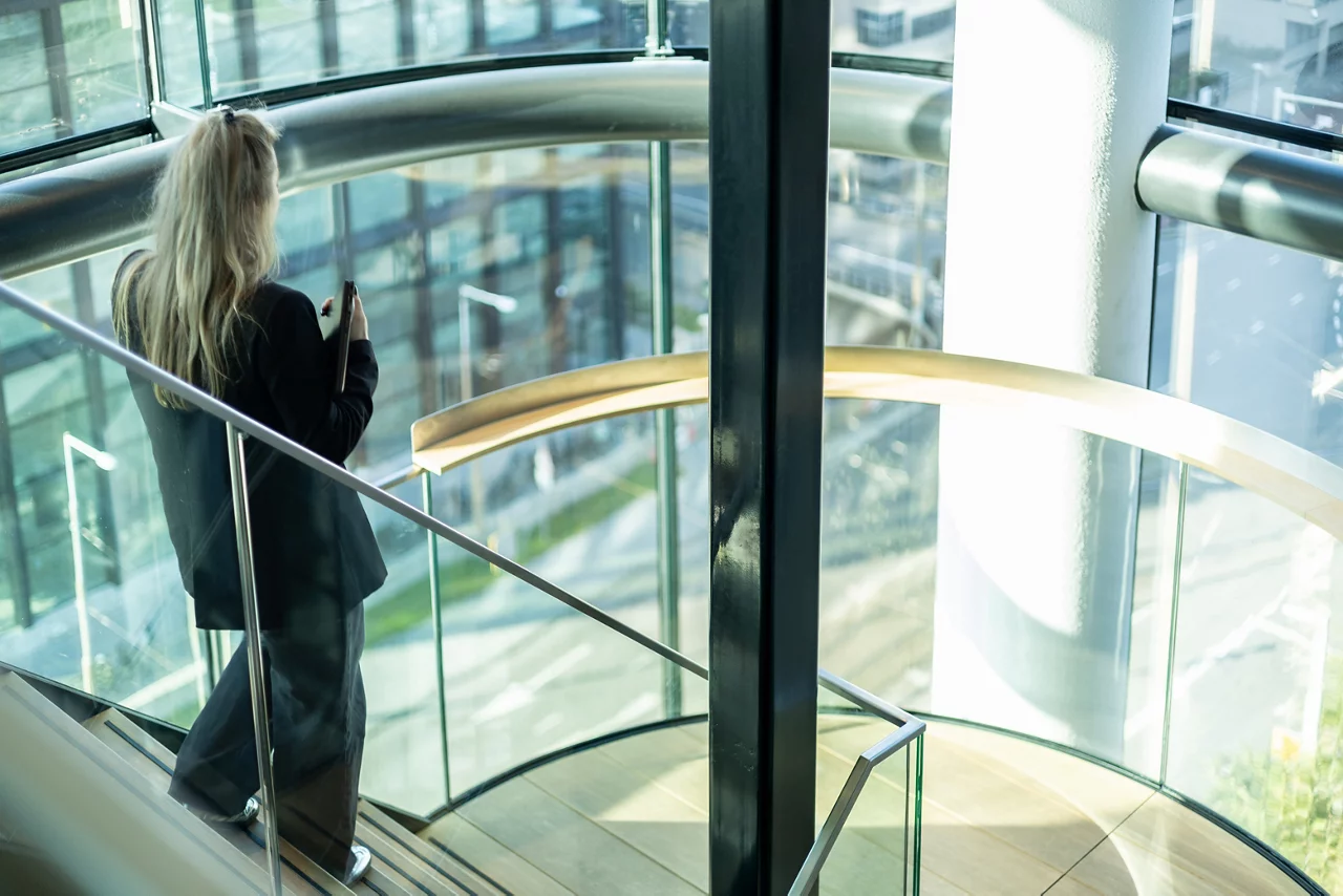 Person in business attire looking out from modern spiral staircase with glass walls overlooking city landscape.