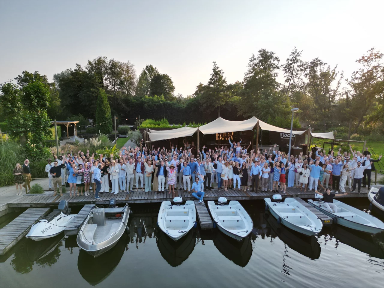 Large group gathered on wooden dock with white boats moored below and event tent in background surrounded by green landscape.