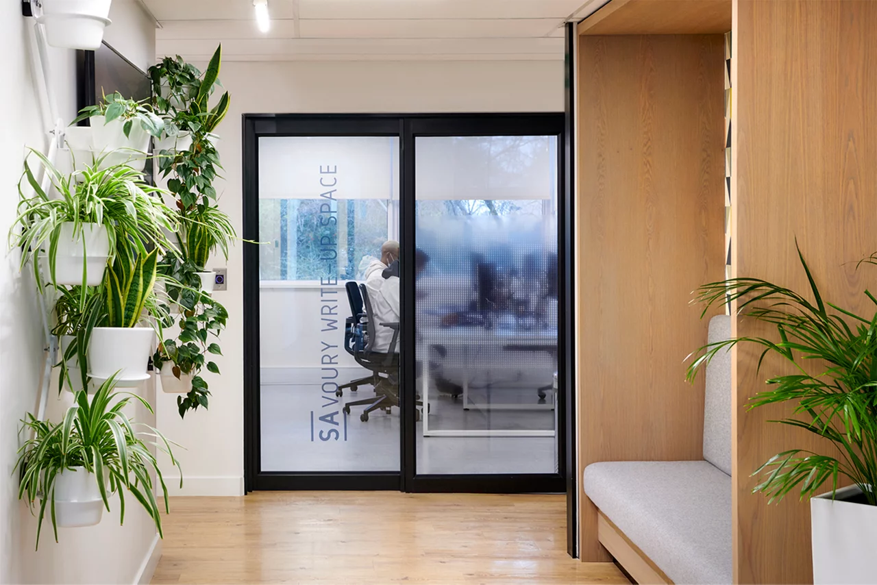 Modern office interior with glass door labeled "Savoury Write Up Space", potted plants, and wooden accents.