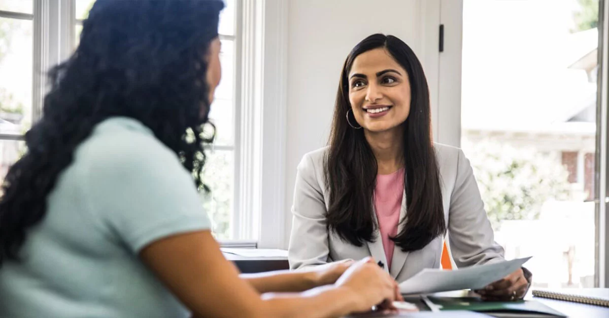 Two professional women engage in a conversation while sitting together in a modern open office space