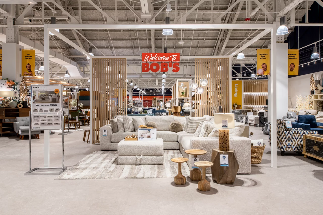 Interior of Bob's furniture store showing a modern display area with light-colored sectional sofa, wooden accent tables, and decorative pillows under a welcome sign