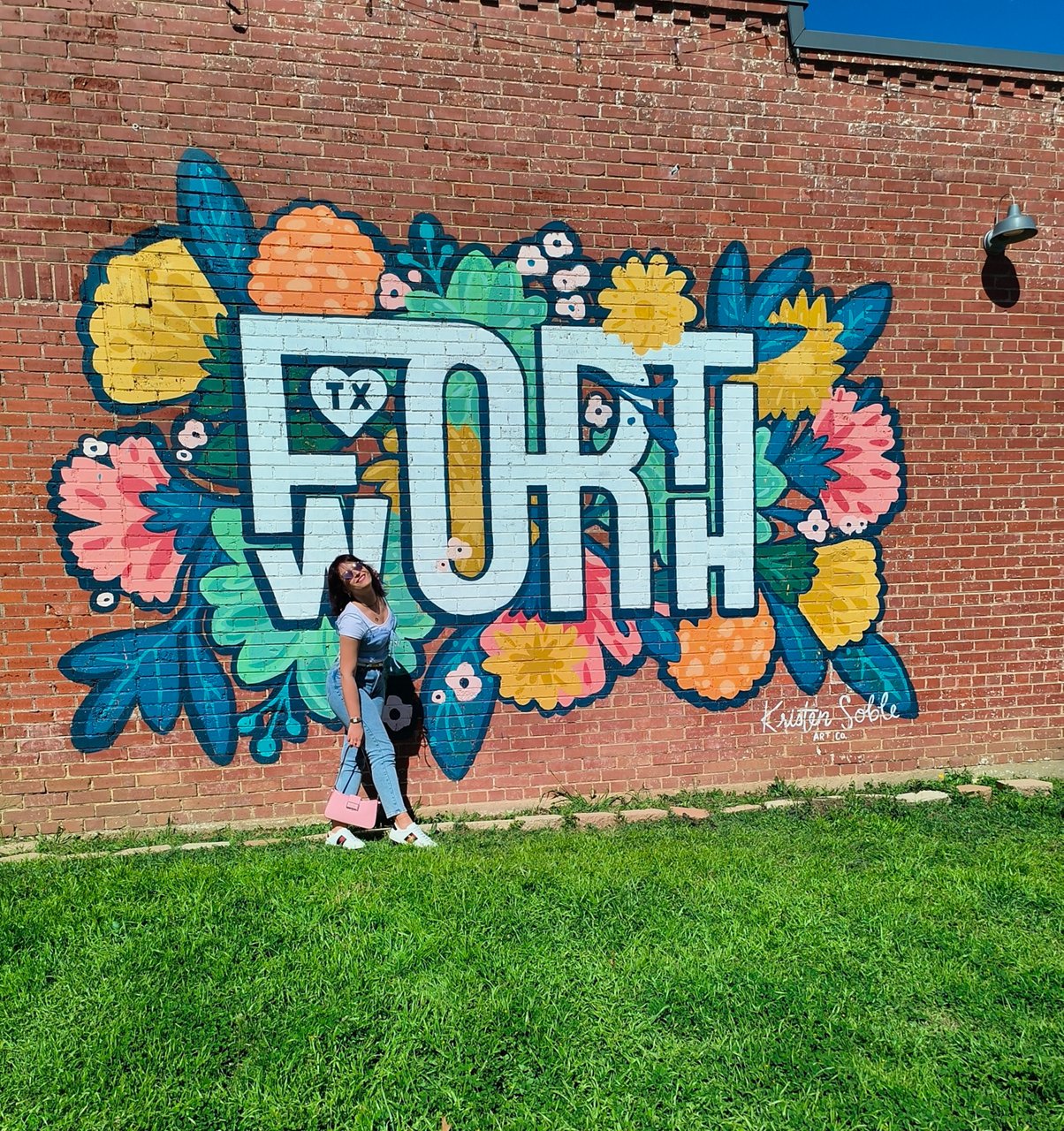  A woman poses in front of a vibrant floral mural on a red brick wall that features large white letters spelling "FORT WORTH"