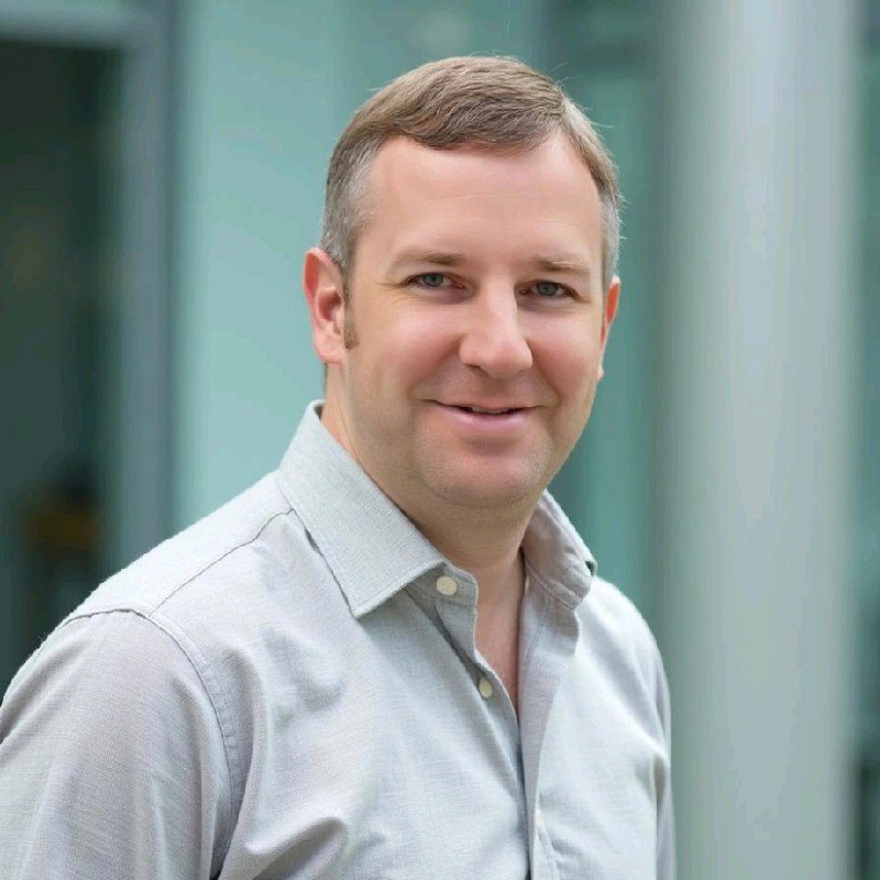 Professional headshot of smiling businessman in light gray button-down shirt against modern office building background.
