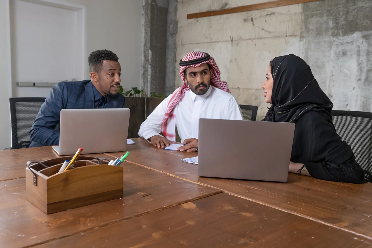 Three diverse business professionals collaborating with laptops at a wooden conference table in a modern office setting.