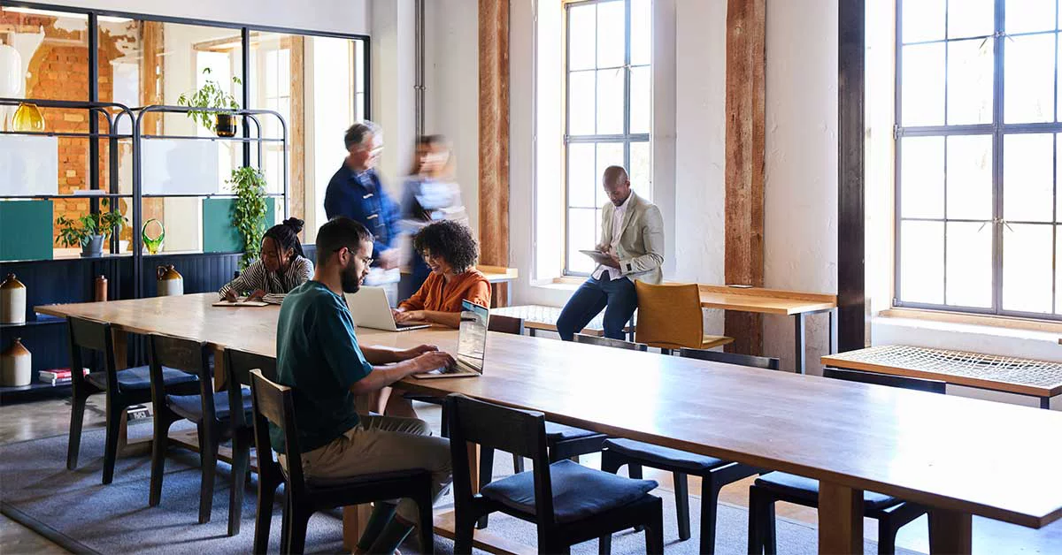  People working at tables in a bright office space with large windows, creating a collaborative environment.