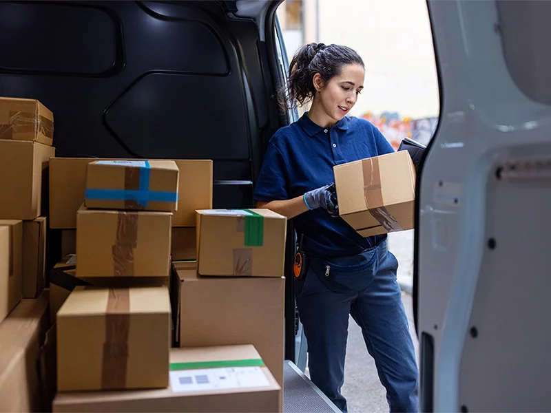 A woman is loading cardboard boxes into the back of a van on a sunny day.