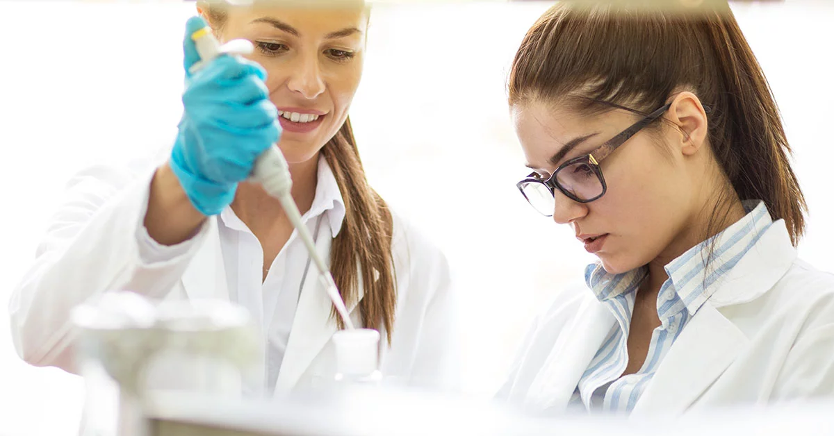 Two scientists in lab coats conducting research, one using pipette while other observes, focused on their work.