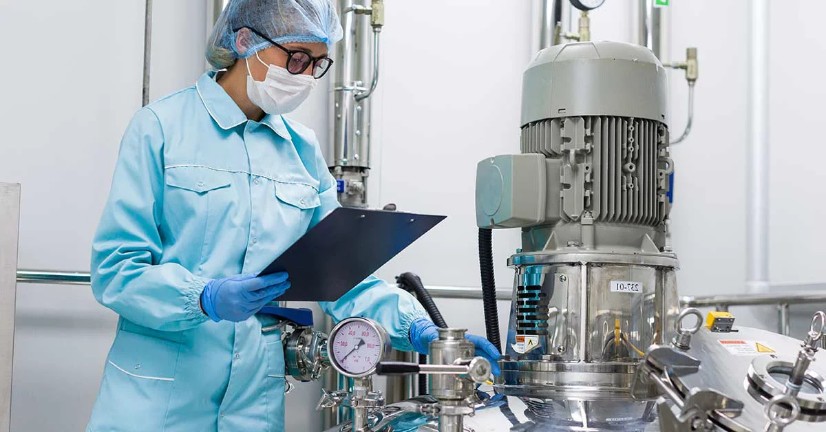 Scientist in protective gear examining equipment in sterile lab, holding clipboard near large stainless steel machinery.