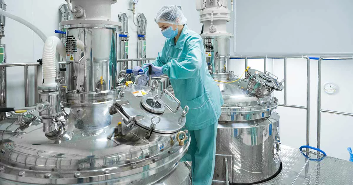 Lab technician in protective gear operating large stainless steel equipment in sterile pharmaceutical manufacturing facility.