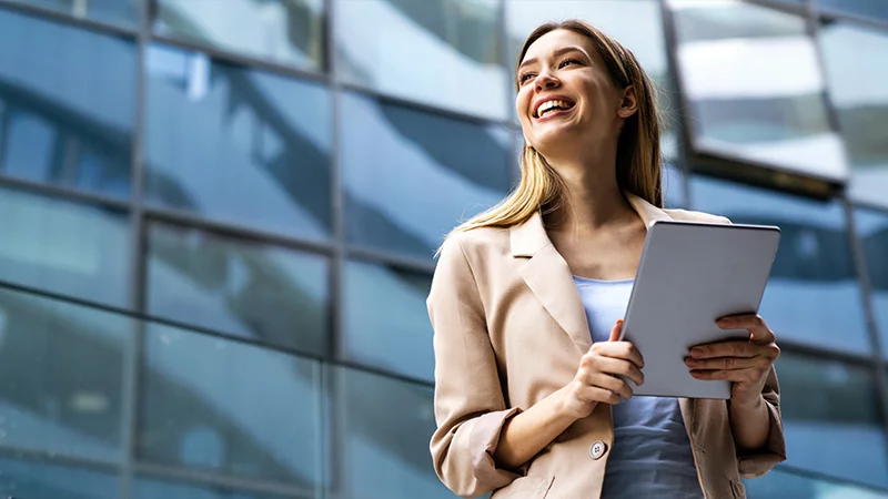 A professional woman in a business suit stands confidently, holding a tablet in her hands.