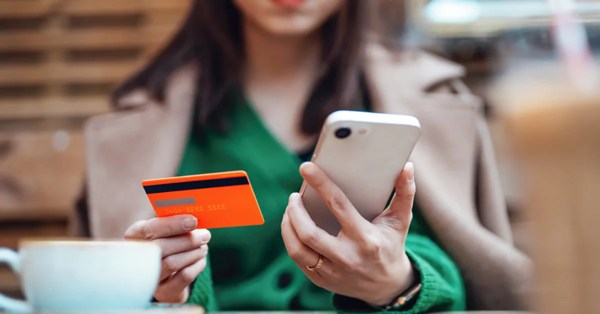 A woman holds a credit card in one hand and a smartphone in the other, appearing focused on a transaction.