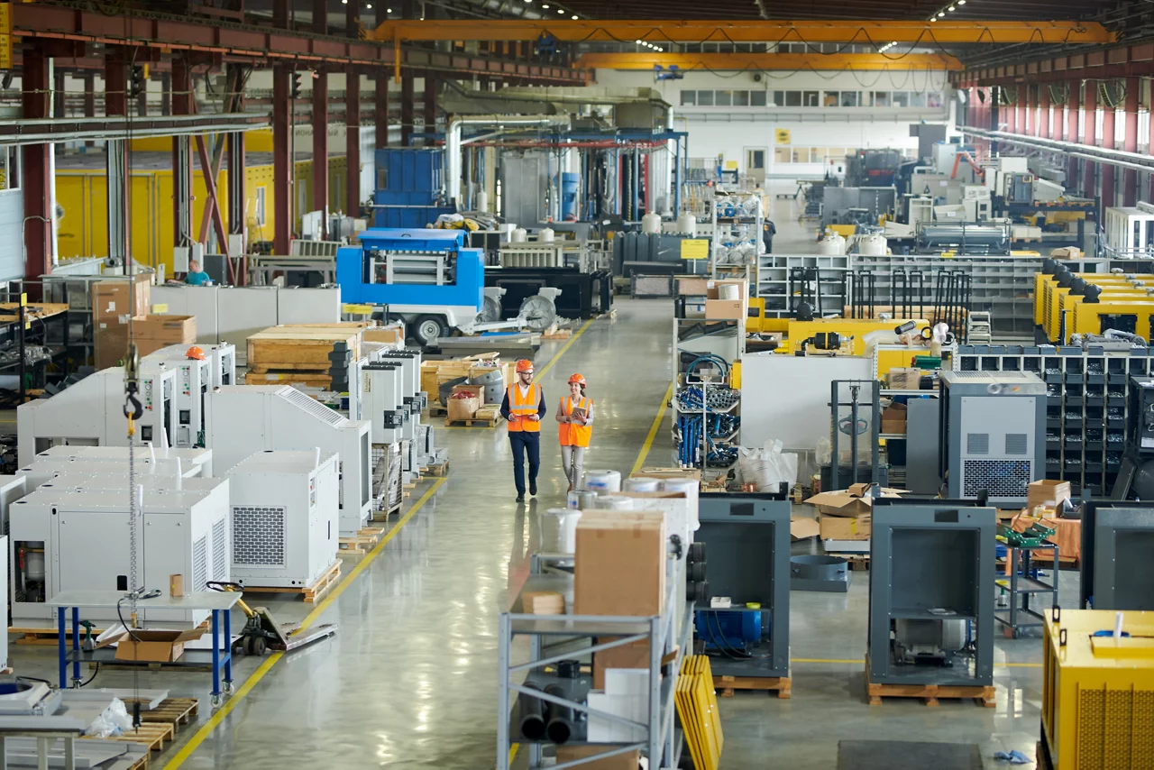 Bearded businessman wearing hardhat walking across production workshop accompanied by female factory employee